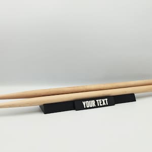 Op de afbeelding: Twee lichtgekleurde houten drumstokken rusten horizontaal op een zwarte rechthoekige displaystandaard. De voorkant van de standaard is voorzien van de witte tekst YOUR TEXT in een vetgedrukt, schreefloos lettertype.