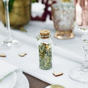 Puede incluir: Una peque&ntilde;a botella de vidrio con un tap&oacute;n de corcho llena de hierbas y flores secas. La botella est&aacute; sobre un mantel blanco con una forma de coraz&oacute;n de madera al lado.