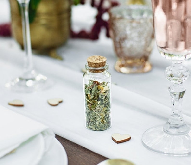 Puede incluir: Una peque&ntilde;a botella de vidrio con un tap&oacute;n de corcho llena de hierbas y flores secas. La botella est&aacute; sobre un mantel blanco con una forma de coraz&oacute;n de madera al lado.
