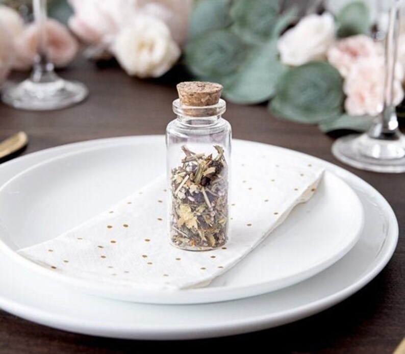 Puede incluir: Una peque&ntilde;a botella de vidrio transparente con un tap&oacute;n de corcho llena de hierbas y flores secas. La botella est&aacute; sobre una servilleta blanca con lunares dorados en un plato blanco.
