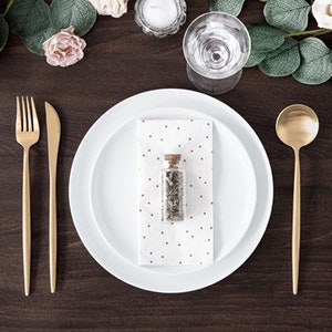 Puede incluir: Un plato blanco con borde dorado y una servilleta blanca con lunares dorados. Una peque&ntilde;a botella de vidrio con tap&oacute;n de corcho y hierbas secas en su interior se coloca sobre la servilleta. Un tenedor, cuchillo y cuchara dorados se colocan sobre una mesa de madera marr&oacute;n oscuro.