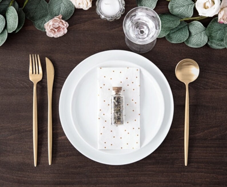 Puede incluir: Un plato blanco con borde dorado y una servilleta blanca con lunares dorados. Una peque&ntilde;a botella de vidrio con tap&oacute;n de corcho y hierbas secas en su interior se coloca sobre la servilleta. Un tenedor, cuchillo y cuchara dorados se colocan sobre una mesa de madera marr&oacute;n oscuro.