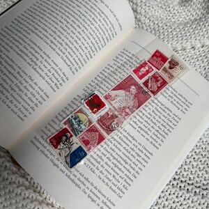 Könnte beinhalten: Ein offenes Buch mit einem Lesezeichen aus alten Briefmarken. Die Briefmarken sind rot, blau und weiß und sind diagonal über die Seite angeordnet. Das Buch liegt auf einer weißen, strukturierten Oberfläche.
