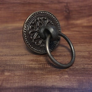 Könnte beinhalten: Ein schwarzer Metall-Ringzug-Schrankknauf mit einem verzierten Design. Der Ringzug ist an einer runden Basis mit einem dekorativen Muster befestigt.