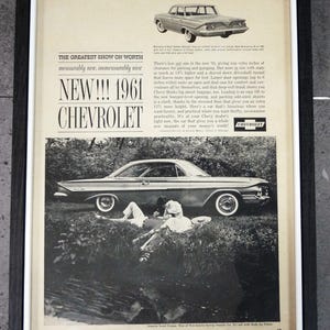 May include: A framed vintage 1961 Chevrolet advertisement featuring black and white photography of a car and a couple resting in grass. The text reads THE GREATEST SHOW ON WORTH, NEW!!! 1961 CHEVROLET, and includes descriptive copy about the vehicle.
