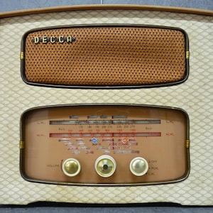 May include: A vintage Decca radio featuring a cream-coloured casing with a diamond-patterned fabric front. The unit includes a brown mesh speaker grille, a glass tuning dial with frequency markings, and three circular control knobs for volume and tuning.