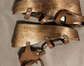May include: Pair of brown leather sandals with wooden platform soles. The sandals feature multiple straps and gold-colored rivet details. The wooden soles have a natural wood grain finish.