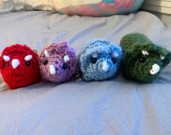 May include: Four crocheted dinosaur keychains in different colors: red, purple, blue, and green. Each keychain features a white horn and black eyes.