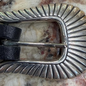 May include: Silver-toned belt buckle with a decorative, fan-like design. The buckle is attached to a black leather belt. The buckle's design features a series of curved, ridged lines radiating from the center, creating a textured appearance.
