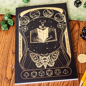 May include: Black notebook with gold foil accents featuring an open book design with a moth and various geometric shapes. The notebook is surrounded by dice and a game map, suggesting a role-playing game theme.