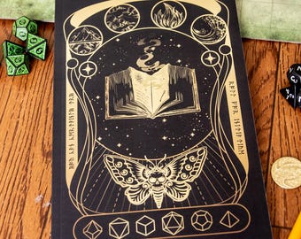 May include: Black notebook with gold foil accents featuring an open book design with a moth and various geometric shapes. The notebook is surrounded by dice and a game map, suggesting a role-playing game theme.