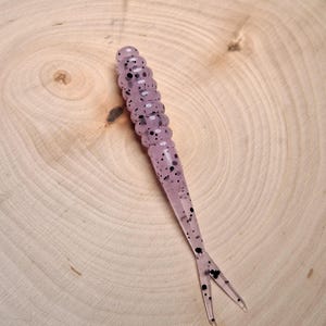 May include: A translucent, lavender-colored fishing lure with black speckles. The lure has a segmented body and a forked tail. The background is a light-colored, wooden surface.