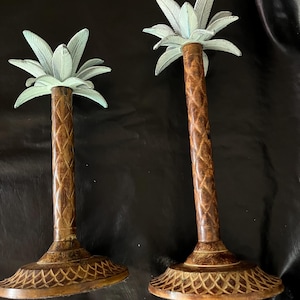May include: Two decorative palm tree candle holders. The palm fronds are a pale green, and the trunks are brown with a textured pattern. The bases are round with a detailed, carved design. The candle holders are made of metal.
