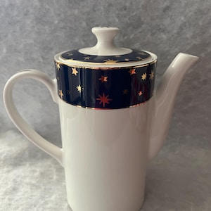 May include: A white porcelain teapot featuring a cylindrical body, a curved handle, and a spout. The lid and the upper section of the teapot are decorated with a deep navy blue band adorned with gold-colored star motifs and thin gold trim.