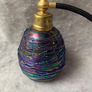 May include: An iridescent perfume atomizer with a rounded glass body featuring a dark purple and green metallic finish with a webbed texture. The bottle is topped with a gold-toned metal cap and a black fabric-covered hose extending to the side.