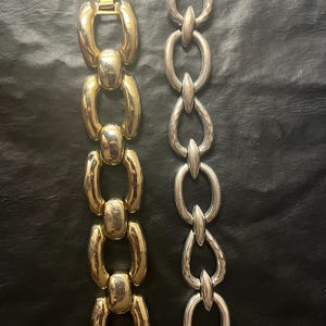 May include: Two chain link bracelets. One is gold-toned with large oval links, and the other is silver-toned with textured oval links. Both bracelets are displayed on a black surface.