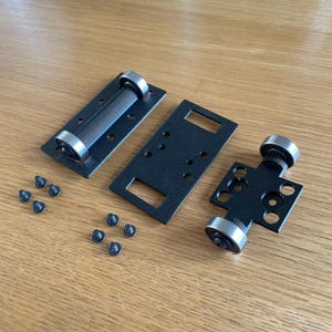 May include: An assortment of black metal hardware components, including plates with rollers and small screws, arranged on a wooden surface. The rollers have silver-colored bearings.