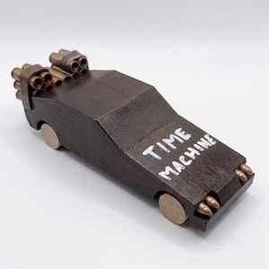 May include: A dark brown wooden toy car with light-colored circular wheels. The hood features the white painted text TIME MACHINE. The rear has two clusters of metallic cylindrical tubes, and the front bumper has four small metallic bullet-like accents.