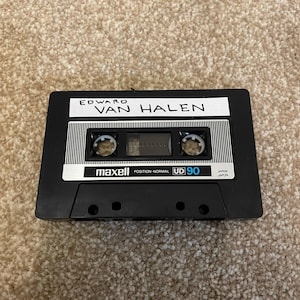 May include: A black Maxell UD 90 audio cassette tape with the band name "Edward Van Halen" printed on a white label. The tape is positioned on a beige carpet.