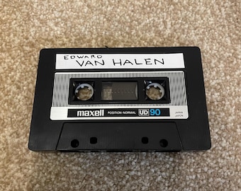 May include: A black Maxell UD 90 audio cassette tape with the band name "Edward Van Halen" printed on a white label. The tape is positioned on a beige carpet.