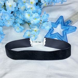 May include: A black velvet choker necklace with a silver clasp. The choker is lying on a white surface with blue flowers in the background.