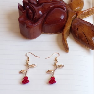 May include: A pair of gold rose earrings with red rose buds. The earrings are on a white surface with a wooden rose sculpture in the background.