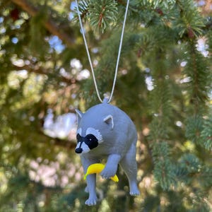 Puede incluir: Un adorno de mapache gris con una m&aacute;scara negra alrededor de los ojos, sosteniendo un peque&ntilde;o pl&aacute;tano amarillo en sus patas delanteras. La figura est&aacute; suspendida por un fino cord&oacute;n plateado, dise&ntilde;ado para colgar de una rama o gancho.