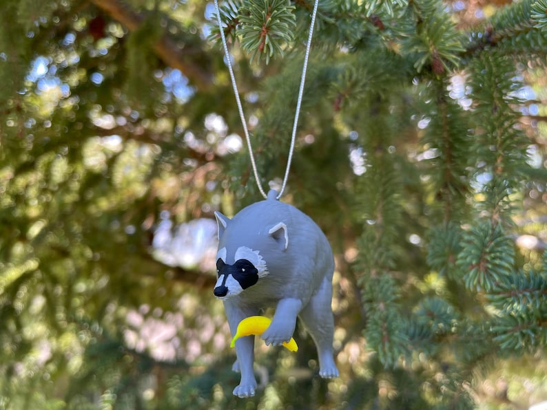 Puede incluir: Un adorno de mapache gris con una m&aacute;scara negra alrededor de los ojos, sosteniendo un peque&ntilde;o pl&aacute;tano amarillo en sus patas delanteras. La figura est&aacute; suspendida por un fino cord&oacute;n plateado, dise&ntilde;ado para colgar de una rama o gancho.