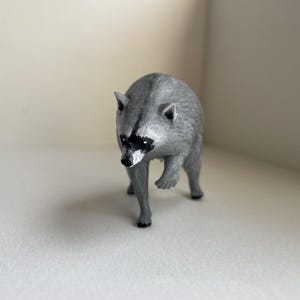 Puede incluir: Una peque&ntilde;a figura de mapache gris con una m&aacute;scara negra alrededor de los ojos y una nariz negra puntiaguda. El animal est&aacute; representado en posici&oacute;n de caminar con una pata delantera ligeramente levantada, con detalles de pelaje texturizado.