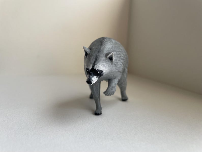 Puede incluir: Una peque&ntilde;a figura de mapache gris con una m&aacute;scara negra alrededor de los ojos y una nariz negra puntiaguda. El animal est&aacute; representado en posici&oacute;n de caminar con una pata delantera ligeramente levantada, con detalles de pelaje texturizado.