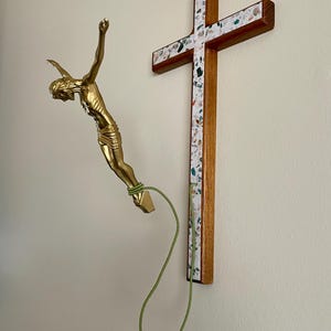May include: A gold-colored sculpture of a figure in a falling pose, connected by a green cord to a wooden cross. The cross has a decorative border with a speckled pattern.