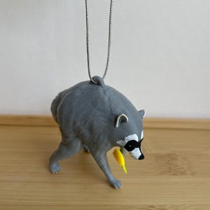 Puede incluir: Un adorno de mapache gris que cuelga de una fina cadena plateada. El mapache se muestra en una postura encorvada con la cabeza hacia abajo, sosteniendo un peque&ntilde;o objeto de color amarillo brillante en su pata delantera.