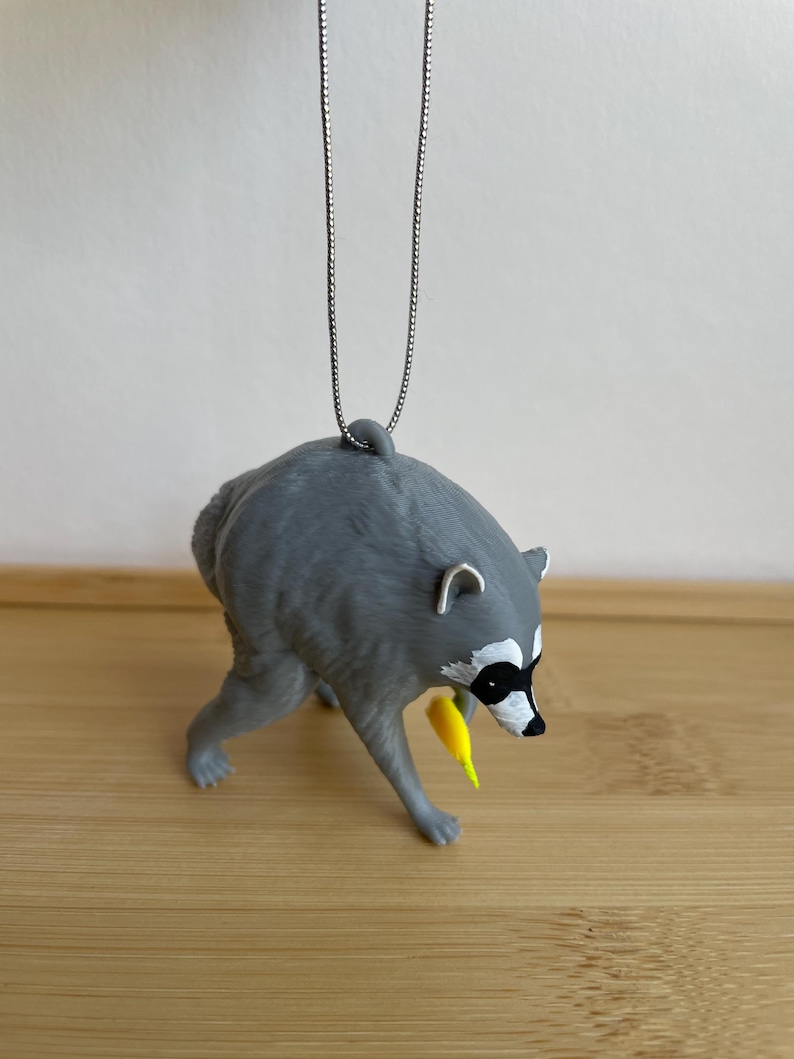 Puede incluir: Un adorno de mapache gris que cuelga de una fina cadena plateada. El mapache se muestra en una postura encorvada con la cabeza hacia abajo, sosteniendo un peque&ntilde;o objeto de color amarillo brillante en su pata delantera.
