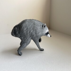 Puede incluir: Una peque&ntilde;a figura de mapache esculpida en color gris, representada en una postura encorvada mientras camina. La figura presenta detalles de pelaje texturizado, un hocico puntiagudo con nariz negra y orejas peque&ntilde;as. Se muestra de perfil.