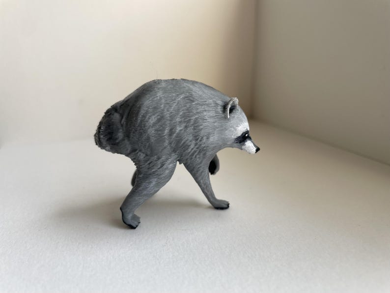 Puede incluir: Una peque&ntilde;a figura de mapache esculpida en color gris, representada en una postura encorvada mientras camina. La figura presenta detalles de pelaje texturizado, un hocico puntiagudo con nariz negra y orejas peque&ntilde;as. Se muestra de perfil.