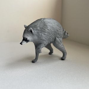Puede incluir: Una peque&ntilde;a figura de pl&aacute;stico gris moldeado de un mapache en posici&oacute;n de caminar. La figura presenta una superficie texturizada que imita el pelaje, un hocico puntiagudo con nariz negra y las caracter&iacute;sticas marcas de m&aacute;scara negra alrededor de los ojos.