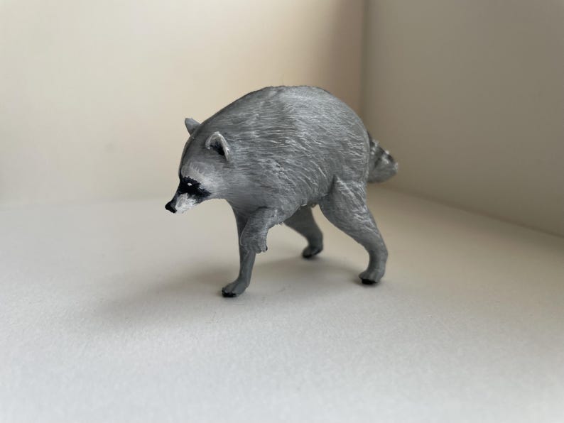 Puede incluir: Una peque&ntilde;a figura de pl&aacute;stico gris moldeado de un mapache en posici&oacute;n de caminar. La figura presenta una superficie texturizada que imita el pelaje, un hocico puntiagudo con nariz negra y las caracter&iacute;sticas marcas de m&aacute;scara negra alrededor de los ojos.