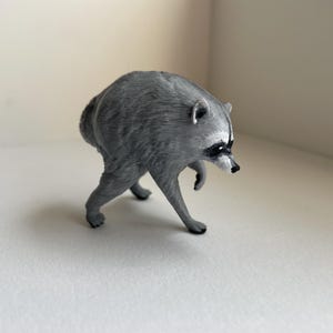 Puede incluir: Una peque&ntilde;a figura de mapache esculpida en color gris, representada en una postura encorvada mientras camina. La figura presenta el caracter&iacute;stico antifaz negro, un hocico puntiagudo con nariz negra y detalles de pelaje texturizado.