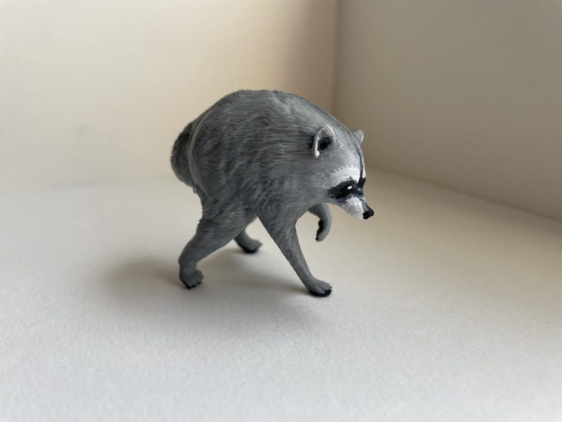 Puede incluir: Una peque&ntilde;a figura de mapache esculpida en color gris, representada en una postura encorvada mientras camina. La figura presenta el caracter&iacute;stico antifaz negro, un hocico puntiagudo con nariz negra y detalles de pelaje texturizado.