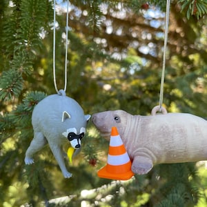 Puede incluir: Dos adornos colgantes que muestran un mapache gris sosteniendo un pl&aacute;tano amarillo y un manat&iacute; de color marr&oacute;n claro junto a un peque&ntilde;o cono de tr&aacute;fico naranja y blanco. Ambas figuras est&aacute;n suspendidas por hilos plateados delgados.