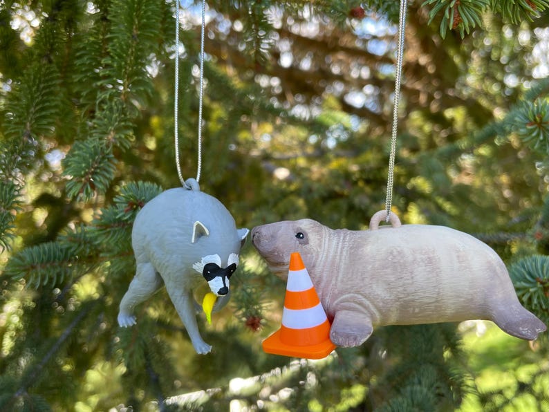 Puede incluir: Dos adornos colgantes que muestran un mapache gris sosteniendo un pl&aacute;tano amarillo y un manat&iacute; de color marr&oacute;n claro junto a un peque&ntilde;o cono de tr&aacute;fico naranja y blanco. Ambas figuras est&aacute;n suspendidas por hilos plateados delgados.