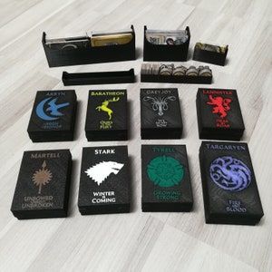 May include: Eight black card boxes with different house sigils from the Game of Thrones TV series. Each box has a different house sigil and motto printed on the front. The houses are: Arryn, Baratheon, Greyjoy, Lannister, Martell, Stark, Tyrell, and Targaryen.
