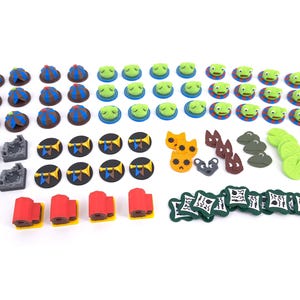 May include: A collection of board game components including small blue and brown tents, green frog figures on blue bases, black circular tokens with yellow trumpets, orange and brown animal shapes, green lily pads, white square tiles, and red buildings.