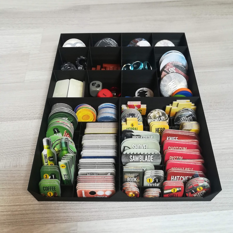 Puede incluir: Un organizador negro con m&uacute;ltiples compartimentos para guardar piezas de juego. Los compartimentos contienen varias cartas, fichas y dados. Las cartas tienen diferentes colores y dise&ntilde;os, incluyendo rojo, verde y amarillo. El texto en las cartas incluye "ALCOHOL", "COFFEE", "HEDDAI", "BROKEN GUITAR", "SHOVEL", "SAWBLADE", "KNIFE", "SHOTGUN", "PISTOL", "BOOK", "SHELL", y "WEAPON".