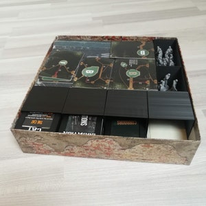 Puede incluir: Un inserto organizador negro para una caja de juego de mesa. El inserto tiene compartimentos para cartas, fichas y miniaturas. Los compartimentos est&aacute;n etiquetados como "Corridors", "Locations", "The Cat" y "Minions".