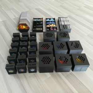 Puede incluir: Un conjunto de cajas de almacenamiento negras con recortes hexagonales, cada una con un color de dados diferente. Las cajas están etiquetadas con nombres como "Saaii", "MINTAK", "CORDURIV" y "NDDIN".