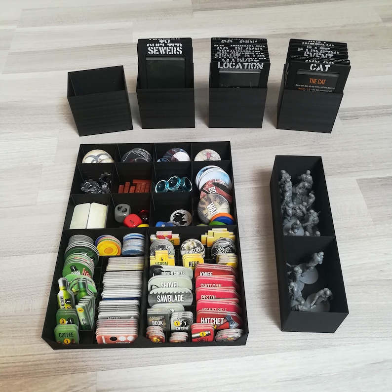 Puede incluir: Un organizador negro con m&uacute;ltiples compartimentos para guardar piezas y cartas de juego. El organizador tiene una etiqueta en blanco y negro que dice "SEWERS". El organizador est&aacute; lleno de varias piezas de juego, incluidas cartas, fichas y dados.