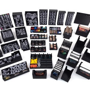 May include: A selection of black storage boxes and trays, filled with miniature figures, tokens, and cards. The figures are various shades of grey, and the cards have colourful illustrations. The boxes are arranged on a white surface.