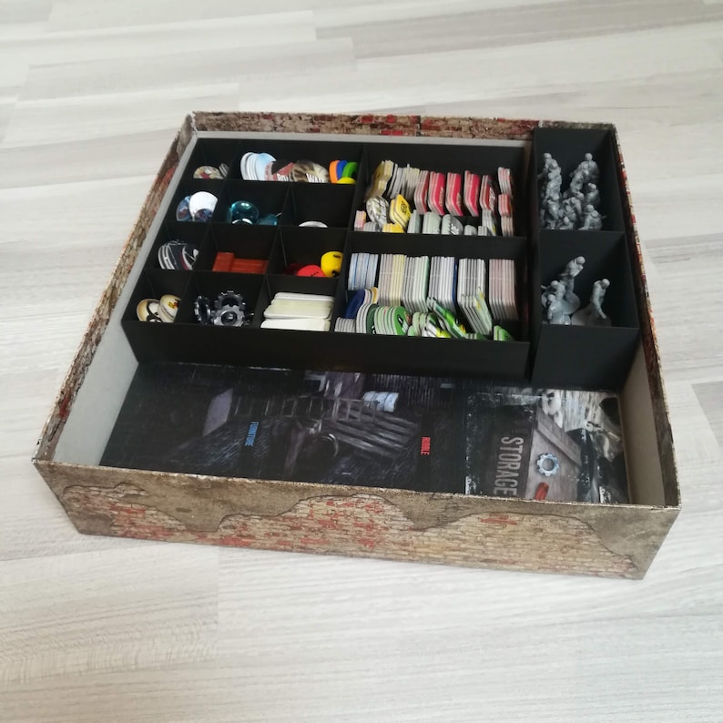 Puede incluir: Una caja de almacenamiento negra con un exterior estampado en marr&oacute;n y rojo. La caja est&aacute; llena de una variedad de piezas de juego, incluyendo cartas, fichas y miniaturas. La caja tiene una etiqueta que dice "STORAGE".