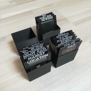 Puede incluir: Dos cajas negras llenas de peque&ntilde;as tarjetas negras. Las tarjetas tienen texto blanco, incluyendo "Night Raid", "Market", "Location", "Culi Tud Sewers", "Little Fate", "Farmers Event", "Color Colt" y "The Cat".